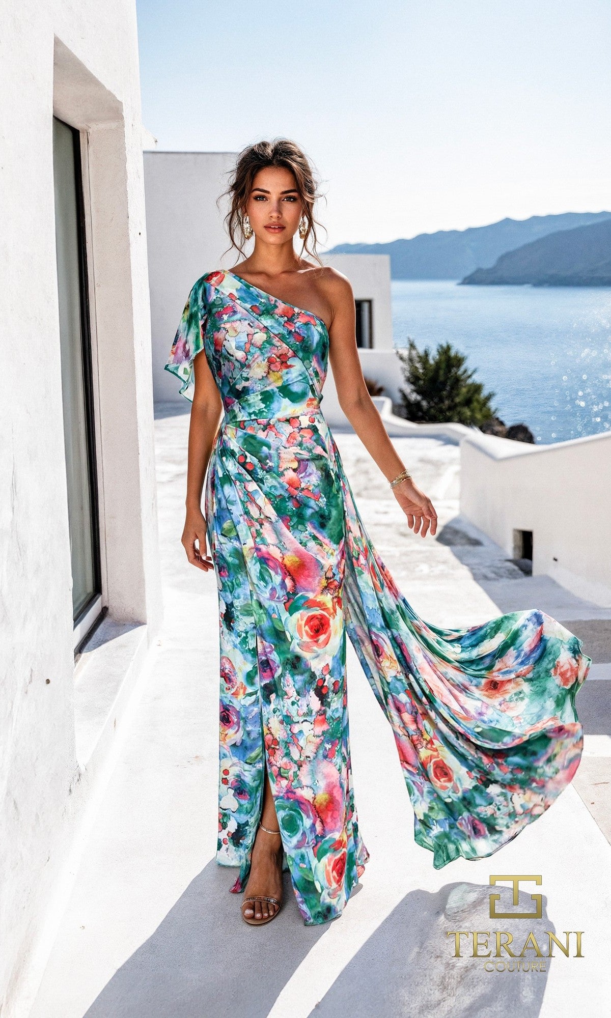 A woman in a green multi Long Formal Dress (Terani 261E6646) stands on a white terrace by the sea, mountains in the background, as her one-shoulder chiffon dress flows elegantly in the breeze.