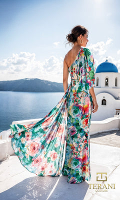 A woman in the Terani 261E6646 Long Formal Dress stands on a white terrace overlooking the sea, with blue domed buildings in the background; the Terani Couture logo is visible.