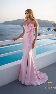 A woman in a pink strapless Terani 261E6670 Mikado formal dress with 3D floral appliques stands on a terrace overlooking a pool and coastal scenery.