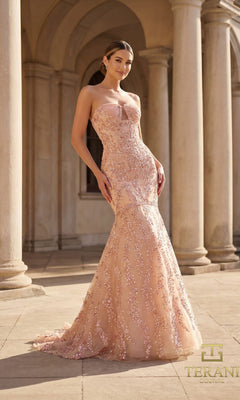 A woman in a blush Terani 261GL6001 long formal dress stands outdoors before stone columns.