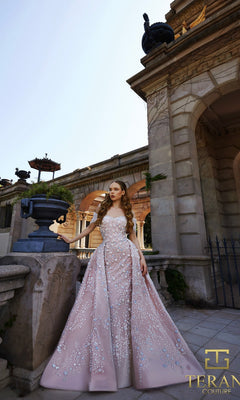 A woman in a pink blue Terani 261GL6659 long formal dress with delicate floral embellishments stands outdoors by a classical stone building, her hand resting on a planter.