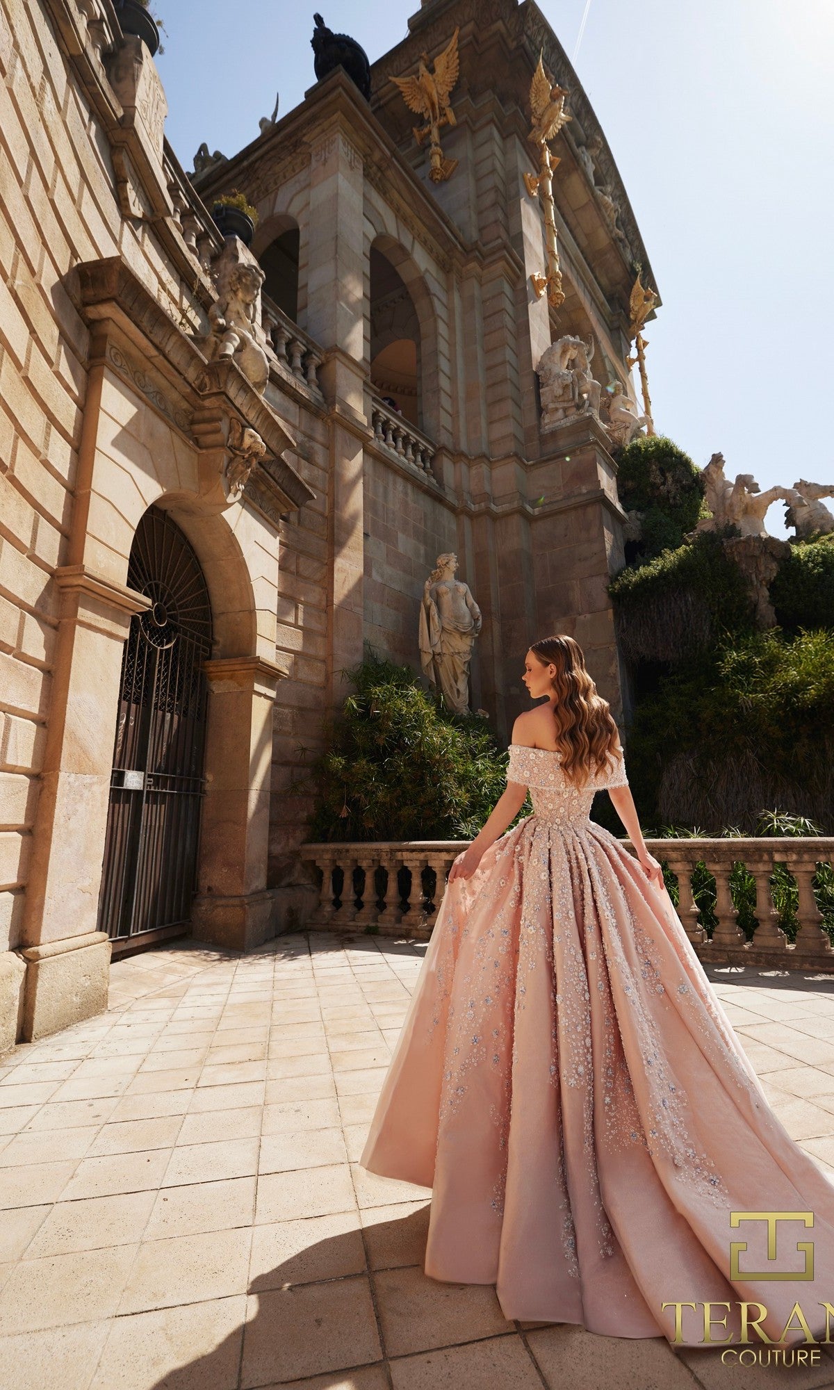 A woman wearing the Long Formal Dress: Terani 261GL6659 stands before a grand stone building decorated with classical statues and intricate architectural details.