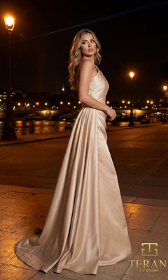 A woman stands on a riverside promenade at night, city lights behind her. She wears a champagne satin Long Formal Dress: Terani 261M6411, its elegant one-shoulder design visible.
