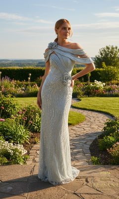 A woman in a sage, off-shoulder Terani 261M6427 gown with floral embellishments and a beaded tulle sheath skirt stands on a stone path in a landscaped garden.