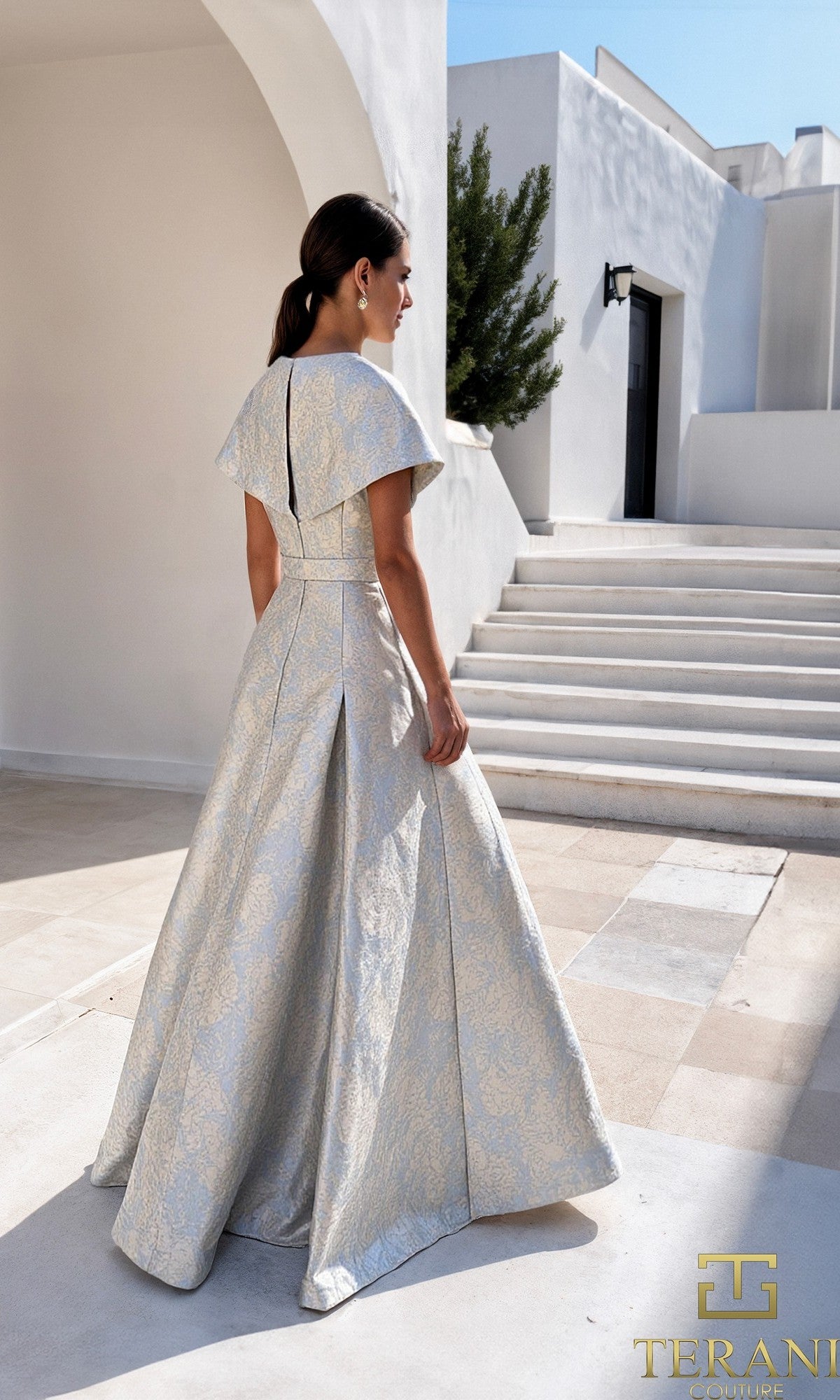 A woman in a Long Formal Dress: Terani 261M6443 stands outdoors on a sunlit patio, with white buildings and stairs in the background.