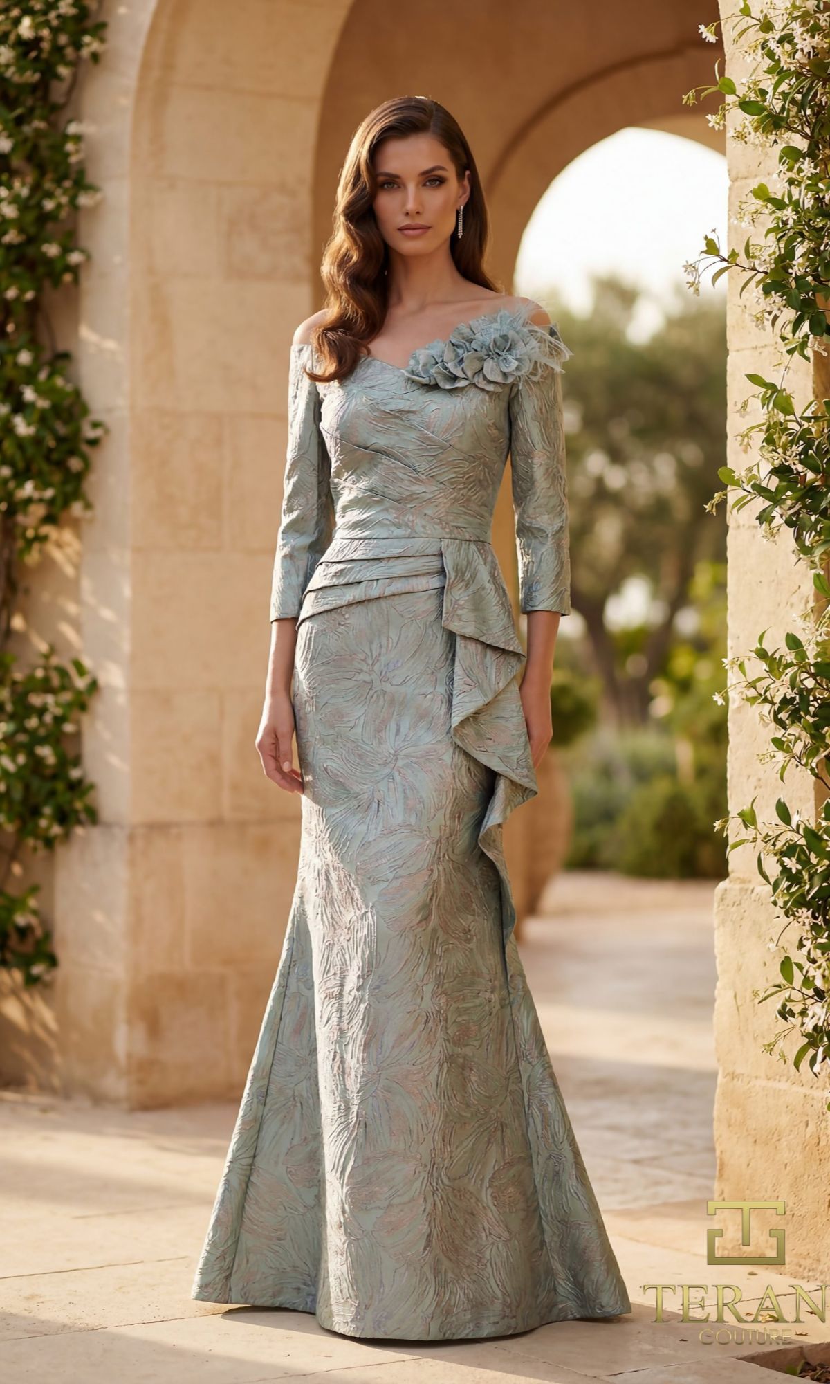 A woman stands outdoors in a light blue Terani 261M6448 long formal dress with an asymmetrical neckline, floral shoulder detail, and ruffled side accent. Stone arch and greenery are visible in the background.