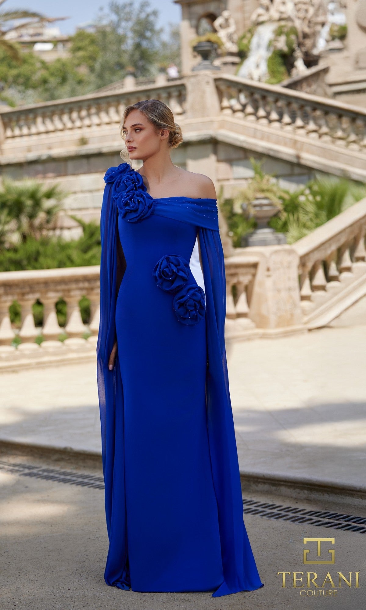 A woman wearing a sapphire Long Formal Dress (Terani 261M6474) with floral accents stands outdoors in front of stone steps and greenery.