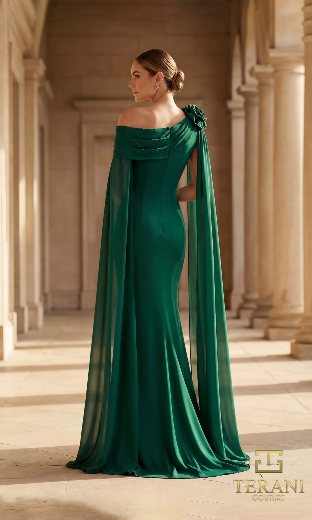 A woman in the Terani 261M6474 long formal dress, featuring an emerald green off-the-shoulder column silhouette, flower shoulder detail, and flowing cape, stands in a classical hallway with columns.