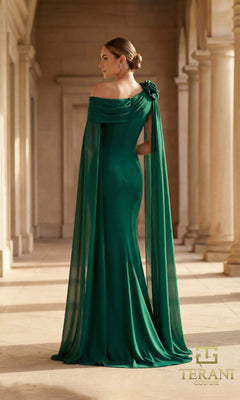 A woman in the Terani 261M6474 long formal dress, featuring an emerald green off-the-shoulder column silhouette, flower shoulder detail, and flowing cape, stands in a classical hallway with columns.