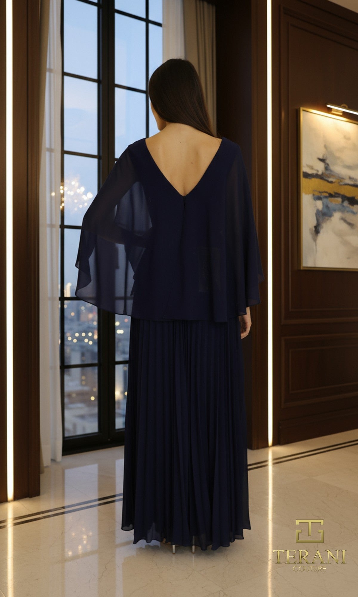 A woman stands indoors near tall windows and elegant wood paneling, wearing the Long Formal Dress: Terani 261M6681 in navy blue, with a pleated chiffon skirt, sheer sleeves, and a deep V back as she faces away from the camera.