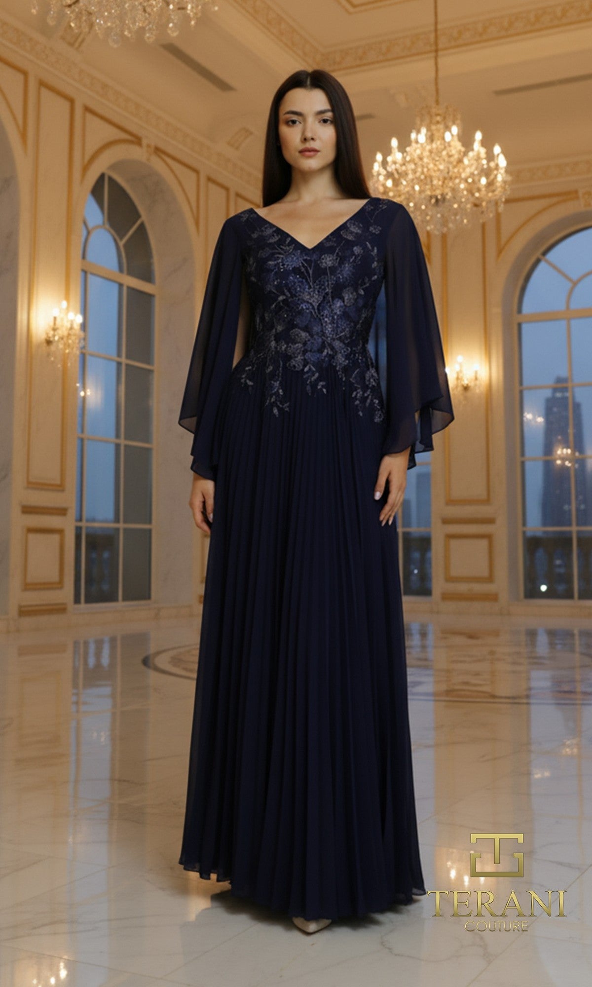 In a marble-floored room, a woman wears the Long Formal Dress: Terani 261M6681 in navy, featuring sheer sleeves, floral embroidery, and a pleated chiffon skirt that flows gracefully beneath elegant chandeliers.
