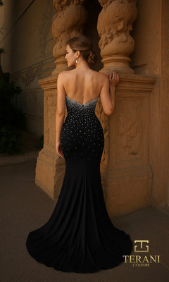 A woman wearing the Terani 261P6030 Long Formal Dress stands with her back to the camera, facing a stone wall. The strapless black gown is fitted with white accents, crystal embellishments, and a slight train.