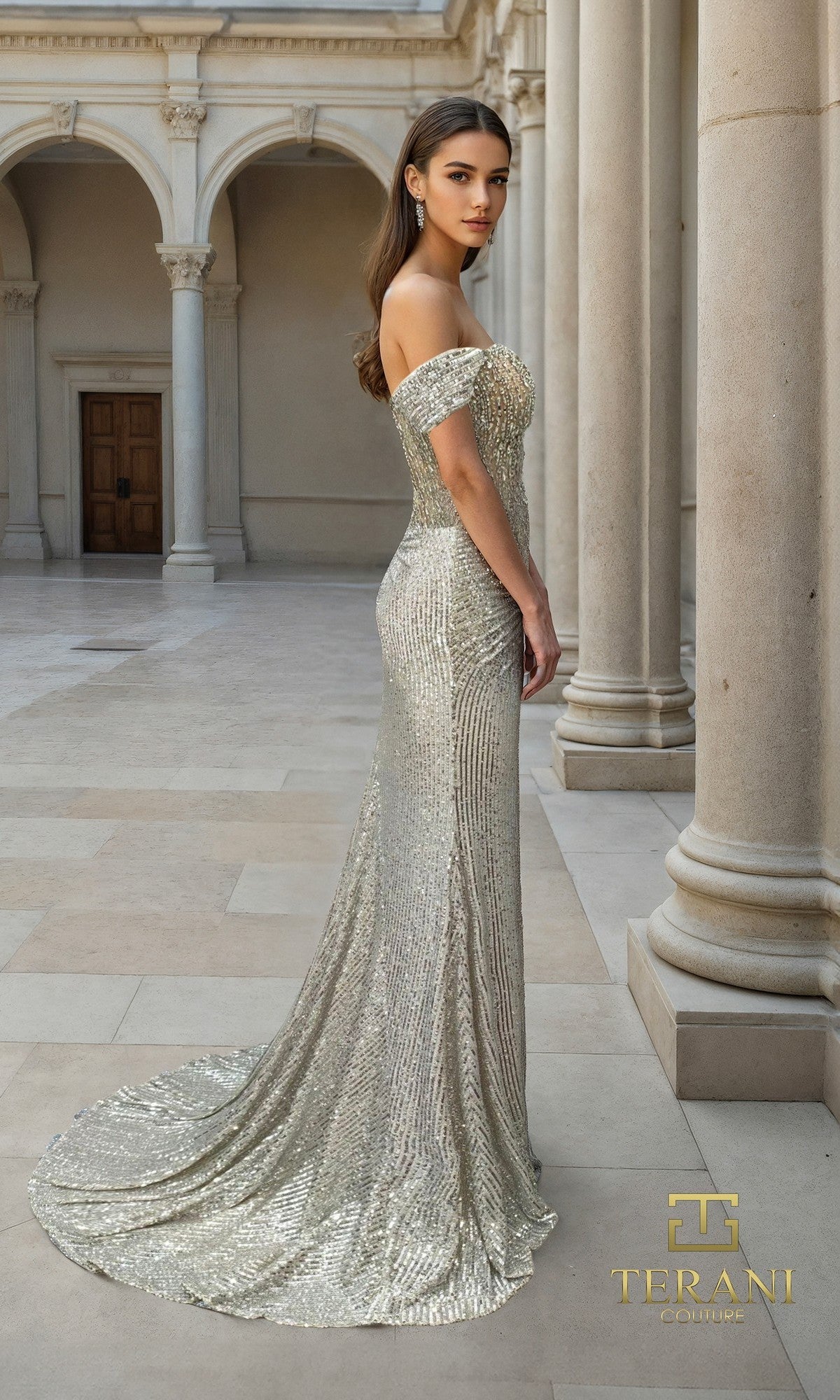 A woman in the Terani 261P6106 long formal dress stands gracefully in a marble courtyard, her off-the-shoulder floor-length silver sequin gown shimmering elegantly among columns and arches.