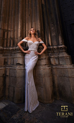 A woman poses against an ornate stone wall, wearing the Terani 261P6161 long formal dress?a white off-the-shoulder gown with intricate embroidered organza patterns. The Terani Couture logo appears in the lower right corner.
