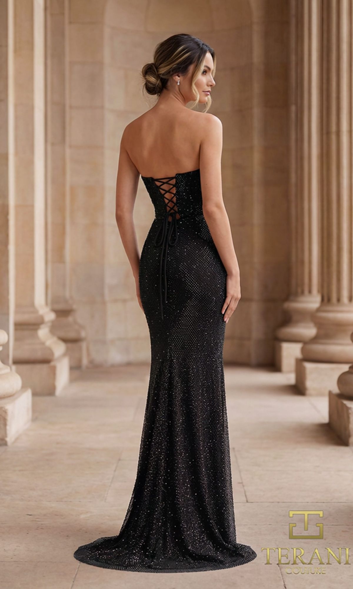 A woman stands on a stone walkway with columns behind her, wearing the Terani 261P6187 long formal dress featuring a strapless corset bodice and lace-up back.