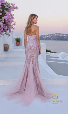 A woman in the Long Formal Dress: Terani 261P6191, a strapless tulle gown, stands on outdoor white steps facing away, with pink flowers and a scenic sunset in the background.