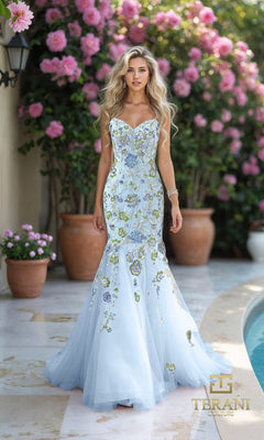 A woman wears the Long Formal Dress: Terani 261P6193 in blue, featuring floral embroidery and a mermaid silhouette, outdoors near potted plants and pink flowers. The Terani Couture logo appears at the bottom right.