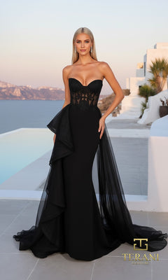 A woman in a strapless black Mikado Long Formal Dress (Terani 261P6261) with sheer panels and a tulle overskirt stands on a terrace overlooking the sea and white buildings; Terani Couture logo is visible.