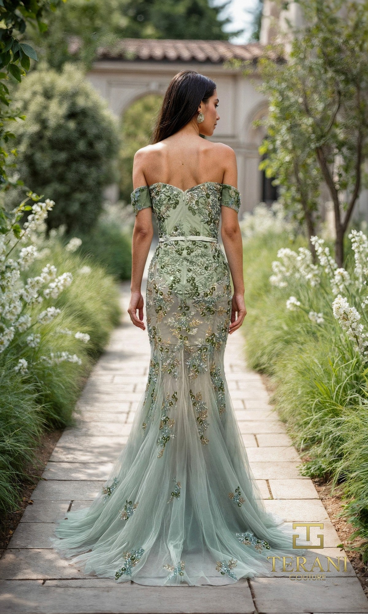 A woman wearing the Terani 261P6676 Long Formal Dress in light green stands on a stone path surrounded by greenery, facing away from the camera.