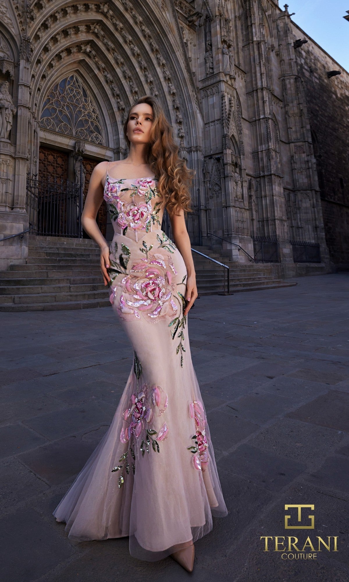 A woman stands before a Gothic cathedral wearing the Terani 261P6677, a fitted sleeveless long formal dress with pink floral embellishments and beading, showcasing elegance and Terani Couture?s signature style.