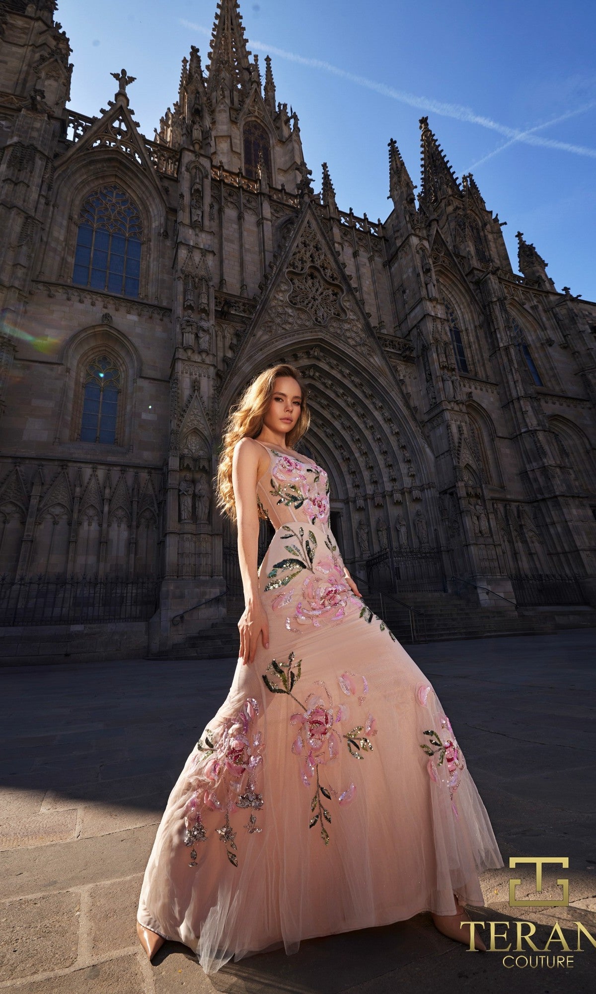 A woman in the Terani 261P6677 long formal dress poses before a Gothic cathedral on a clear day; Terani Couture logo appears in the corner.