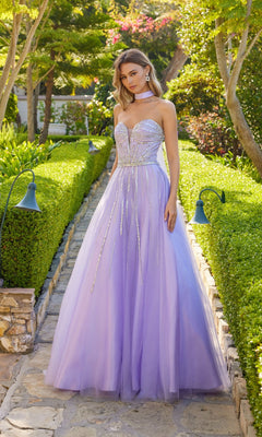 A woman wearing the Long Prom Dress: Juliet JT2620U in lilac, featuring a strapless tulle design and rhinestone-embellished bodice, stands on a stone path in a manicured garden.