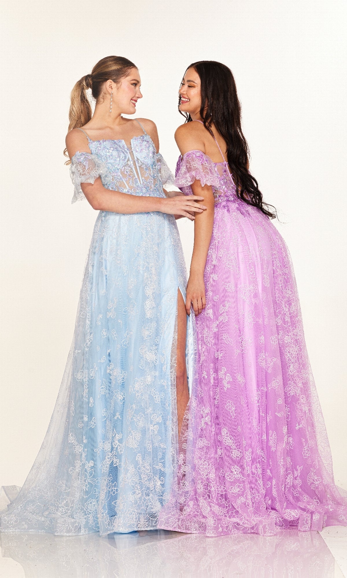 Two women stand together in Cecilia Couture 2620 long prom dresses—one blue, one lavender—with lace details and sheer corset bodices, smiling at each other against a plain background.