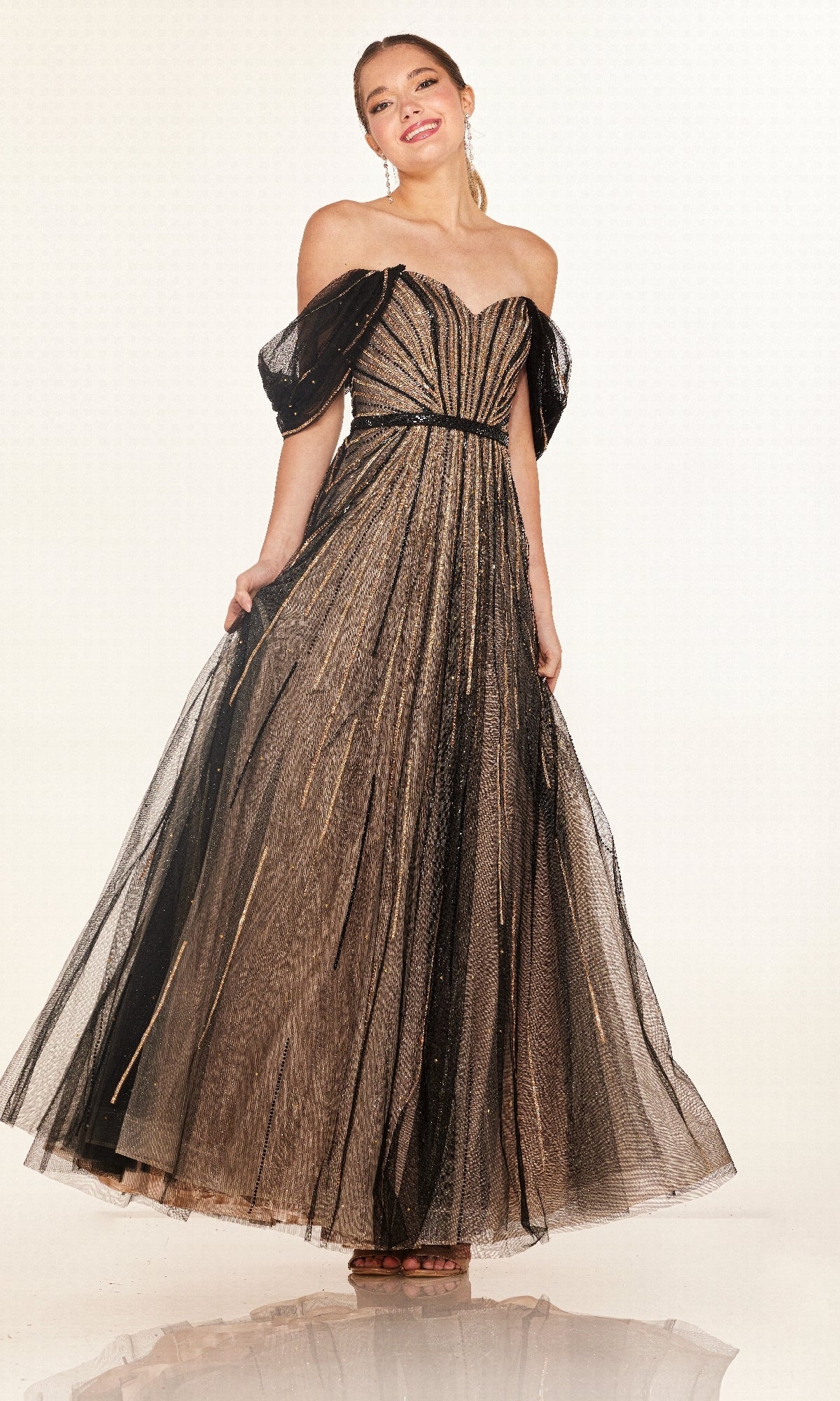 A woman poses and smiles against a plain light background wearing the Cecilia Couture 2621 long prom dress, featuring black and gold sequin embellishments, an off-the-shoulder neckline, and a flowing tulle skirt.