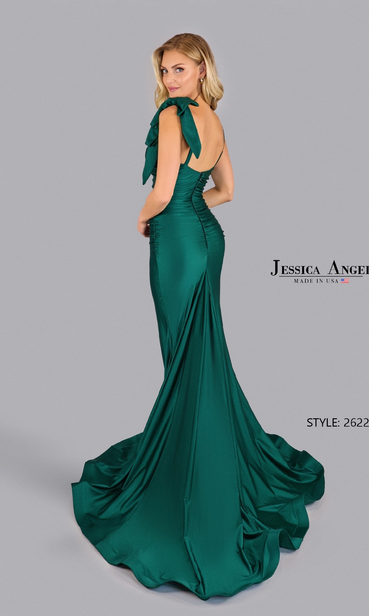 Woman poses with her back to the camera in a floor-length, emerald green Shoulder-Bow Long Prom Dress by Jessica Angel 2622, featuring a bow detail on the shoulder against a plain background.