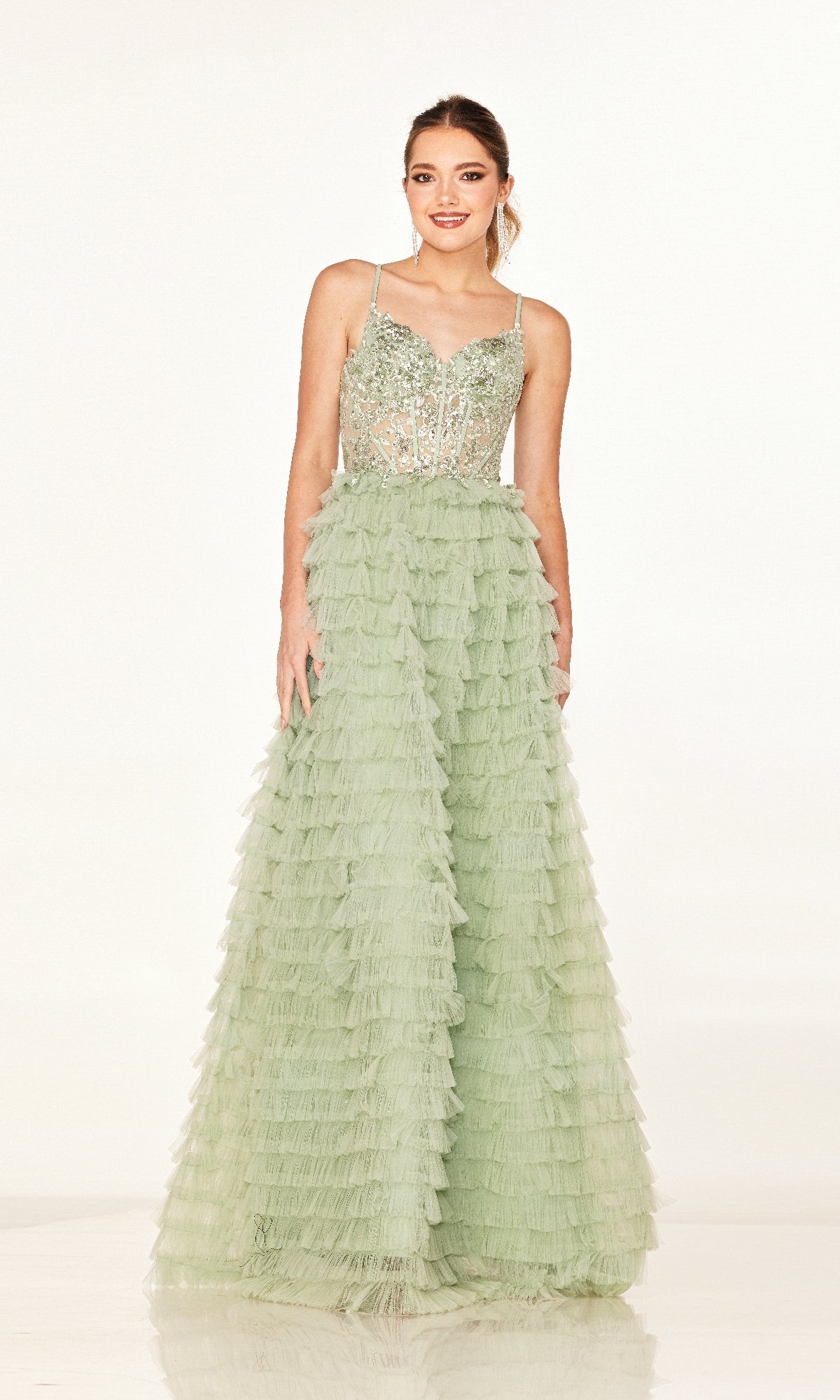 A woman wears the Long Prom Dress: Cecilia Couture 2624—a light green A-line gown featuring a sequined bodice, thin straps, tiered tulle skirt, and lace-up back—while standing against a plain white background.