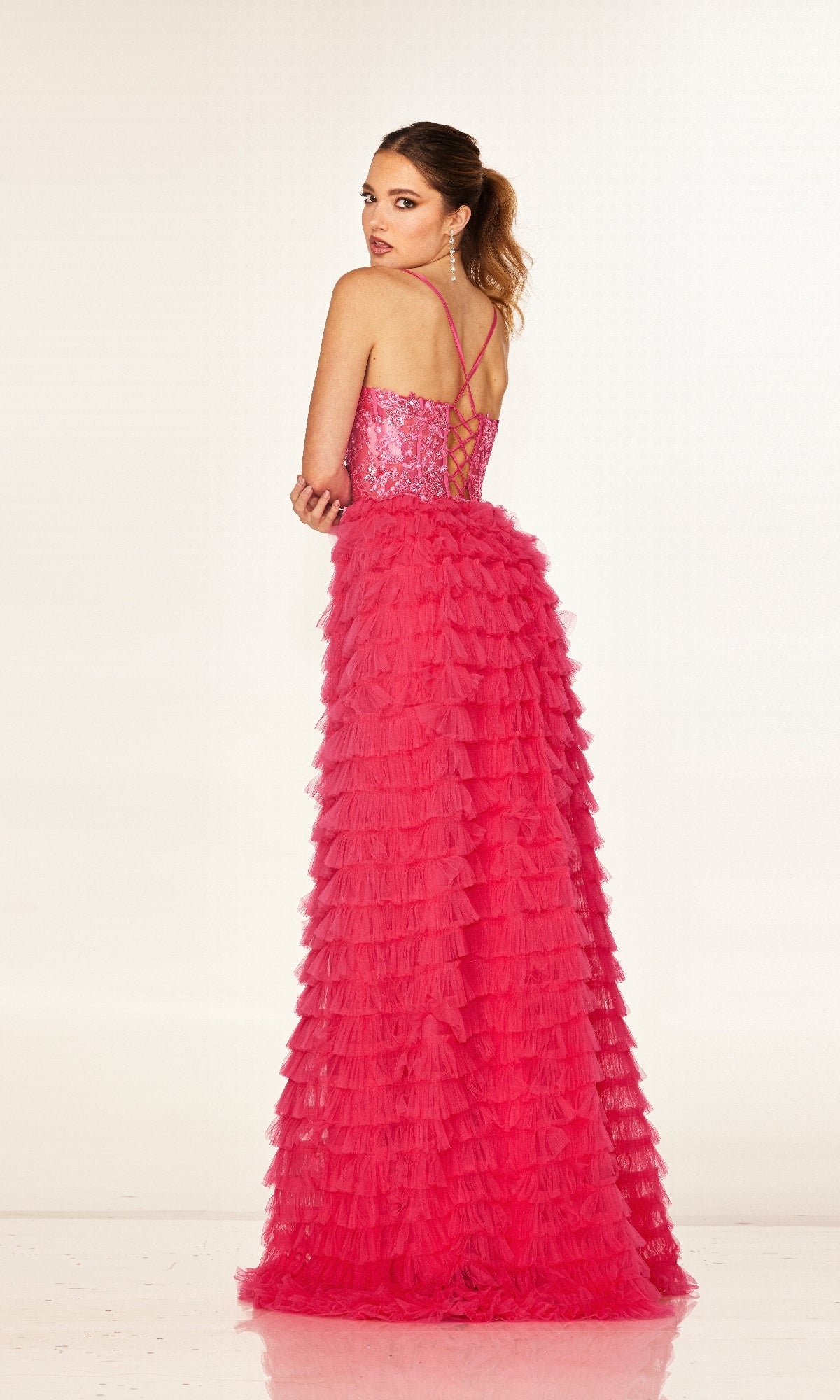 Wearing the Cecilia Couture 2624 long prom dress, a woman in a bright pink, floor-length A-line gown with ruffles, a sequined fitted bodice, and thin crisscross straps stands facing away, glancing over her shoulder.