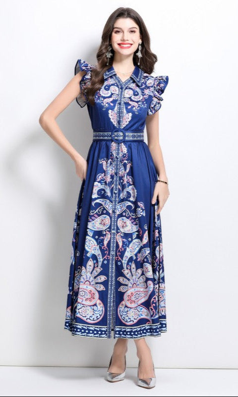 A woman in a navy blue paisley print casual maxi dress with a belted waist and ruffled cap sleeves, paired with silver heels, stands against a plain white background.