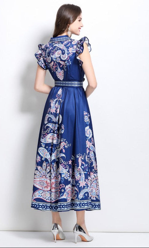 A woman faces away, wearing the Navy Blue Paisley Print Casual Maxi Dress with ruffled cap sleeves and a belted waist, styled with silver heels against a plain white background.