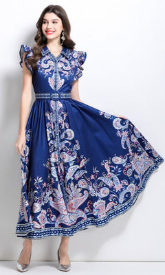 A woman models the Navy Blue Paisley Print Casual Maxi Dress with a belted waist and ruffled cap sleeves, standing against a white background and holding out part of the ankle-length skirt.