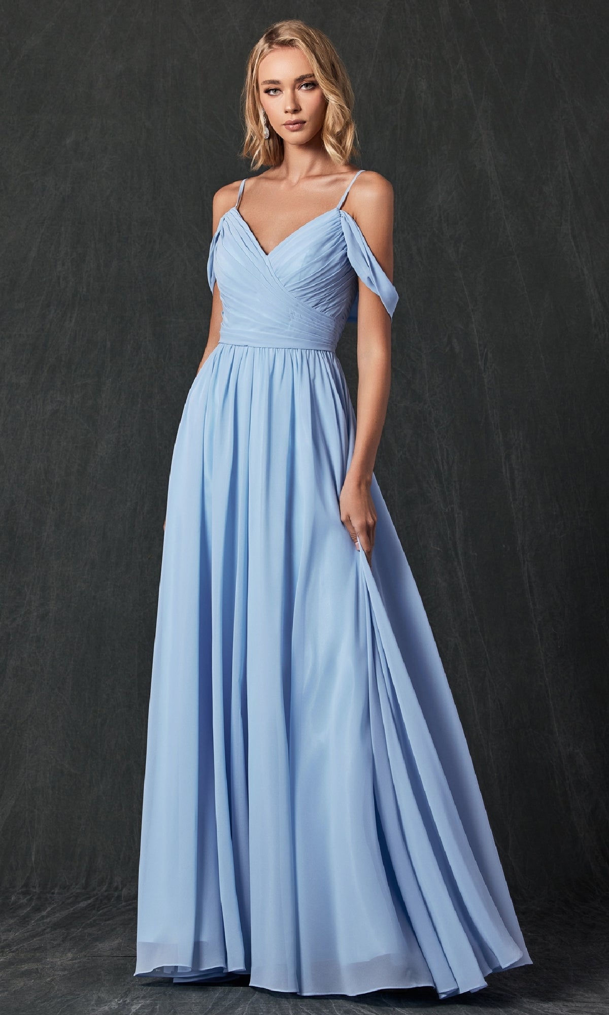 A woman poses against a dark backdrop in the Juliet 262 long prom dress, featuring a light blue, floor-length A-line design with off-the-shoulder straps and a fitted pleated bodice. She gracefully holds part of the dress in one hand.