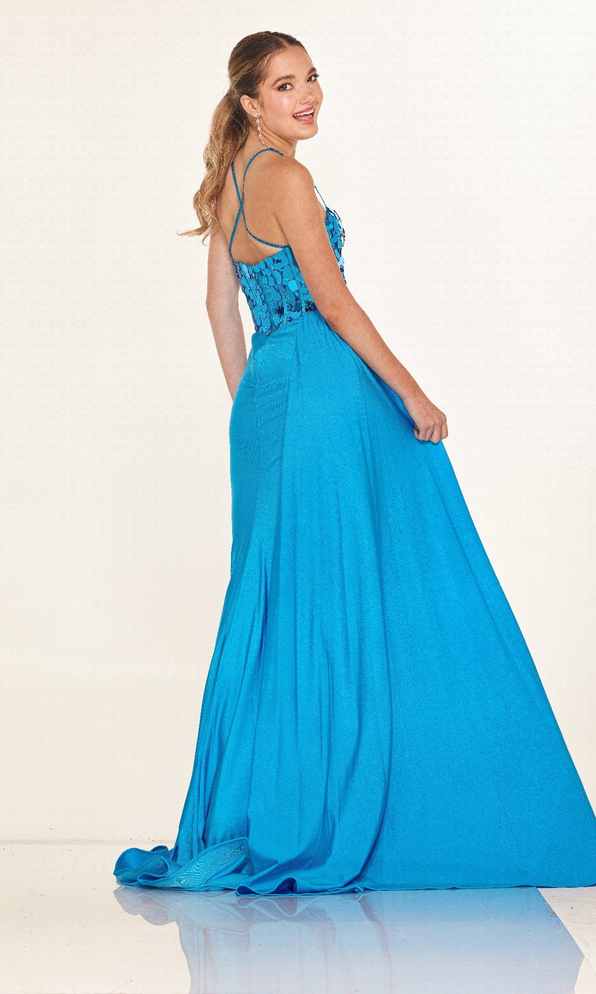 Woman wearing the Cecilia Couture 2630 long blue prom dress with thin straps and a lace bodice, standing and looking over her shoulder against a white background.