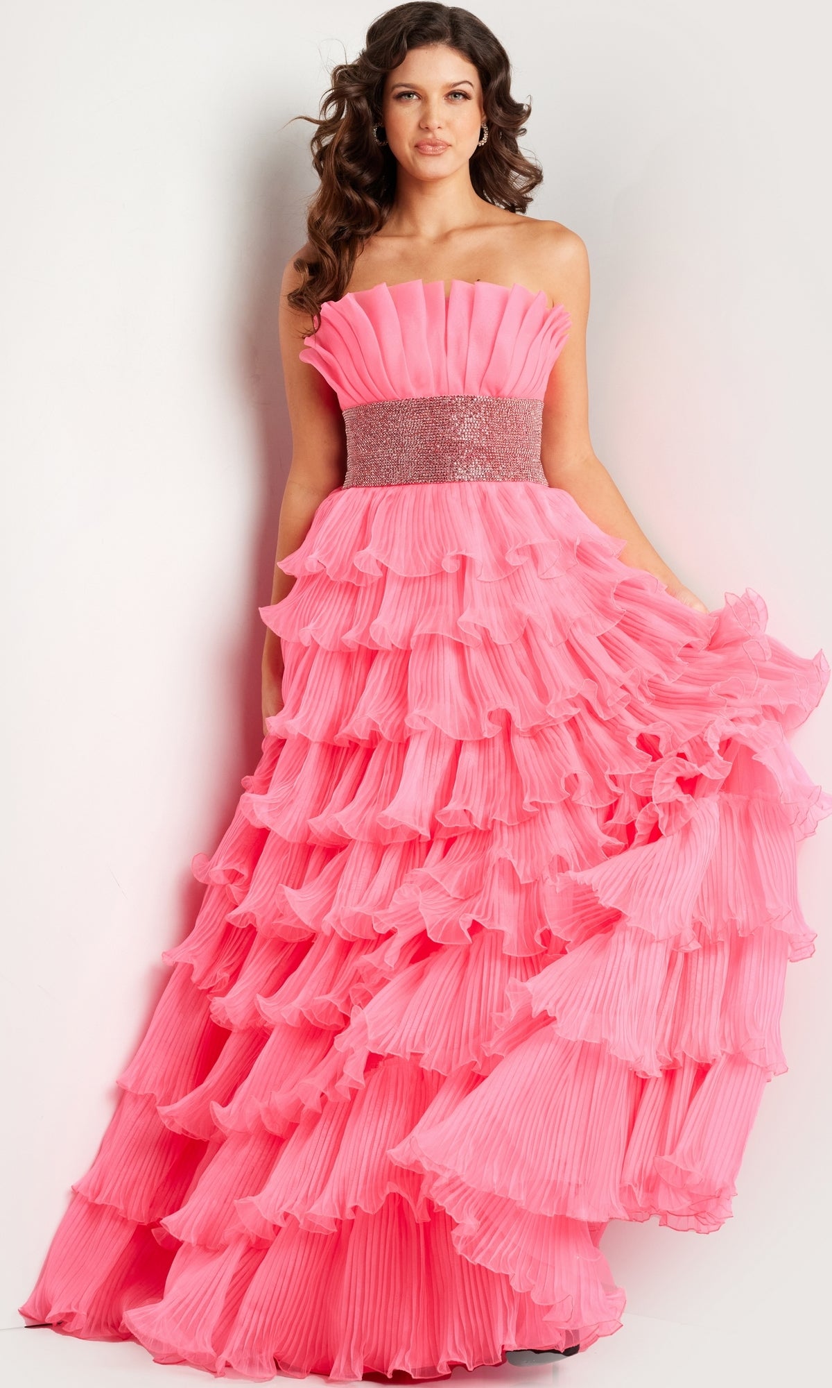 A woman wears the Jovani Strapless Long Tiered Prom Ball Gown 26314, featuring a fitted, beaded waistband and floor-length pink ruffled tiers, standing against a plain background.