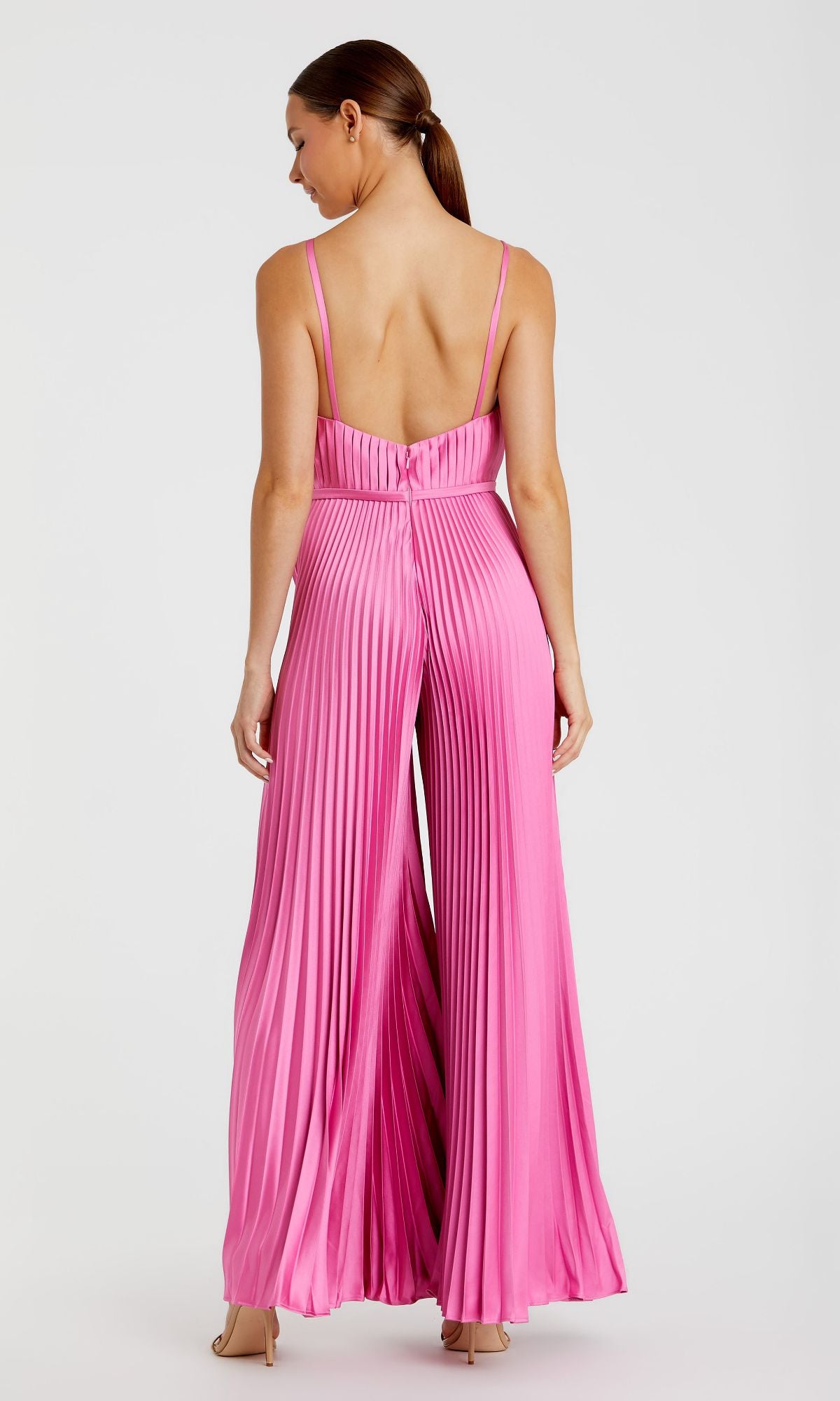 A woman faces away, wearing the Mac Duggal Pleated Wide-Leg Formal Jumpsuit 26319 in pink with a sleeveless, backless design and nude heels, set against a plain light background.