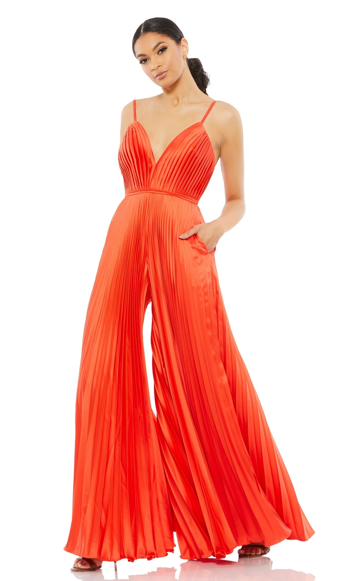 A woman in the Mac Duggal Pleated Wide-Leg Formal Jumpsuit 26319, featuring bright orange color, spaghetti straps, and wide-leg pants, stands against a white background.