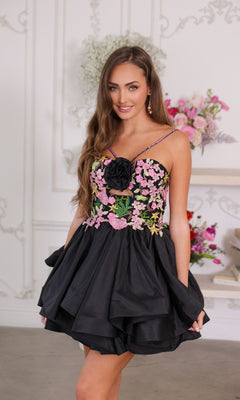 A woman wearing the Ava Presley 26348 short black homecoming dress with pink floral embroidery smiles at the camera indoors, with floral arrangements in the background.