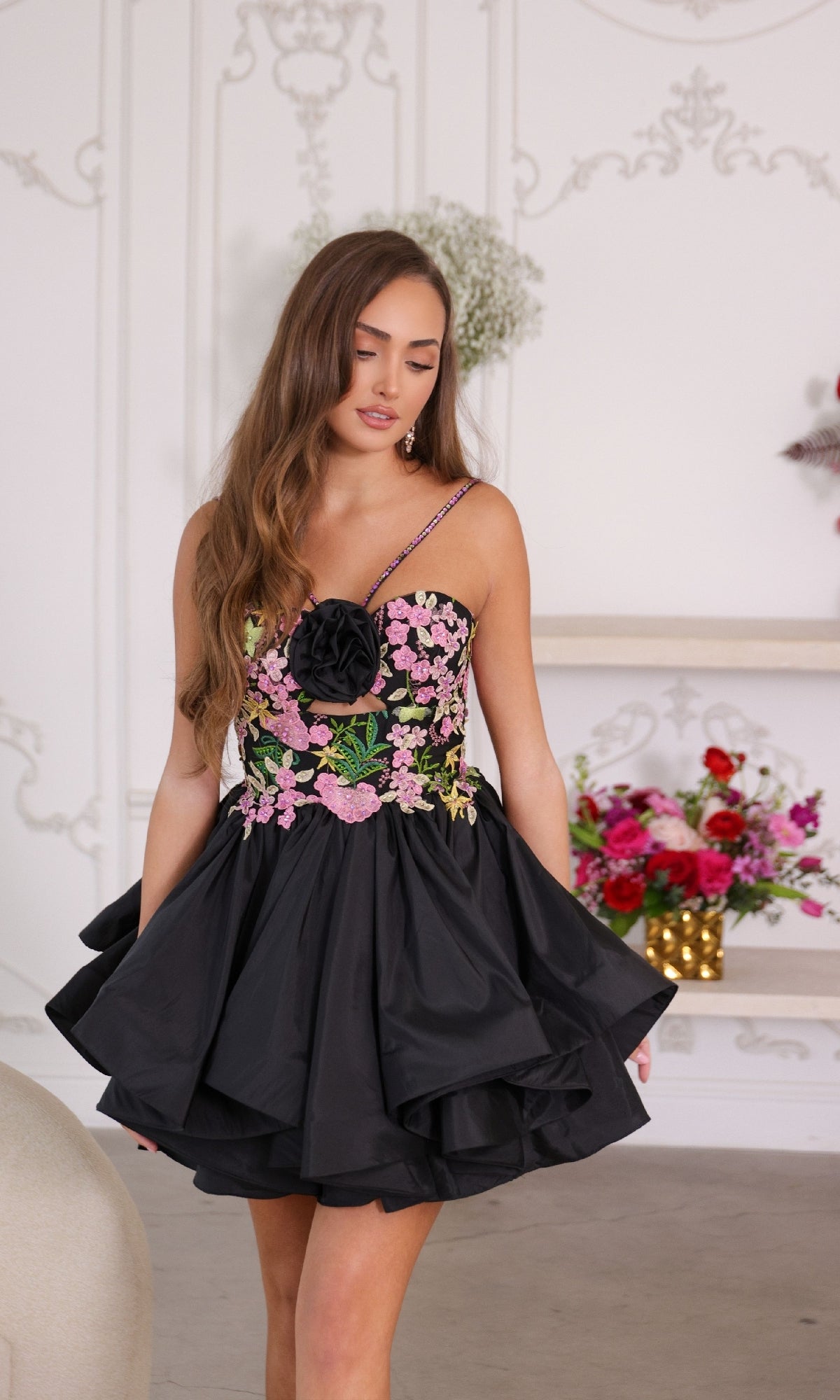 A woman stands indoors among floral arrangements, wearing the Ava Presley 26348 short homecoming dress—a black mini dress with ruffles and pink floral embroidery.