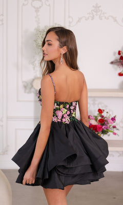 A woman indoors looks over her shoulder wearing the Ava Presley 26348 short homecoming dress, featuring a sleeveless black design with a layered skirt and floral-embroidered bodice. Flowers can be seen in the background.