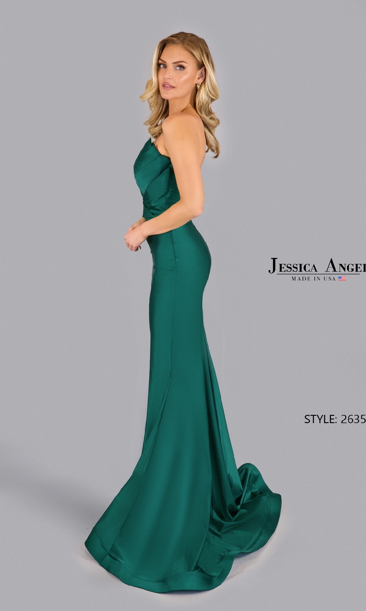 A woman models the Pleated Strapless Prom Gown by Jessica Angel (Style 2635), an emerald green fitted mermaid dress with a slight train, standing against a plain gray background. 