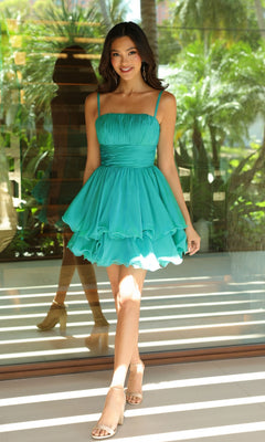 A woman wears the Ava Presley 26378 Tiered Short A-Line Hoco Dress in teal with nude heels, standing indoors with greenery visible through the glass behind her.