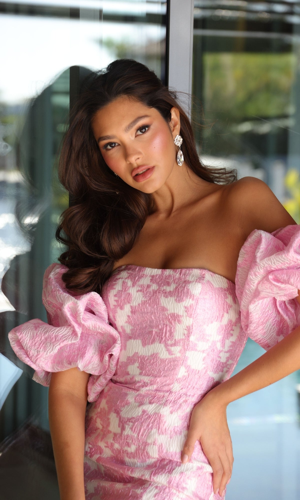 A woman poses indoors in the Short Homecoming Dress: Ava Presley 26391—a pink dress with puffed sleeves—standing in front of glass windows and wearing statement earrings.