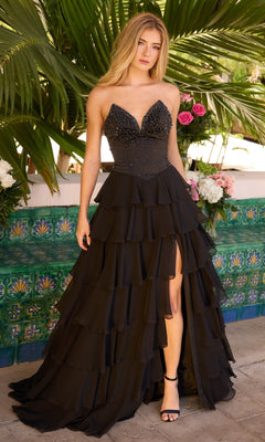 A woman stands outdoors in the black Ava Presley 26407 strapless ruffled prom gown, featuring a beaded bodice and tiered skirt; palm leaves and flowers form a lush backdrop.