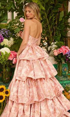 A woman in the Ava Presley 26412 Ruffled Strapless Prom Gown stands among vibrant flowers, holding a pink rose and glancing over her shoulder in her floral print evening gown.