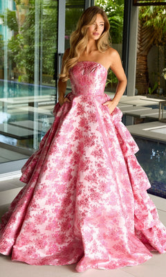 Indoors by a pool, a woman wears the Ruffled Strapless Prom Gown: Ava Presley 26412, an A-line prom dress with pink floral print, full skirt with pockets, and elegant back ruffles.