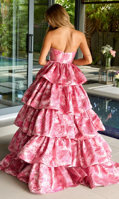 A woman stands indoors by a pool, her back to the camera, wearing the Ava Presley 26412 Ruffled Strapless Prom Gown—a voluminous a-line dress with pink floral print.