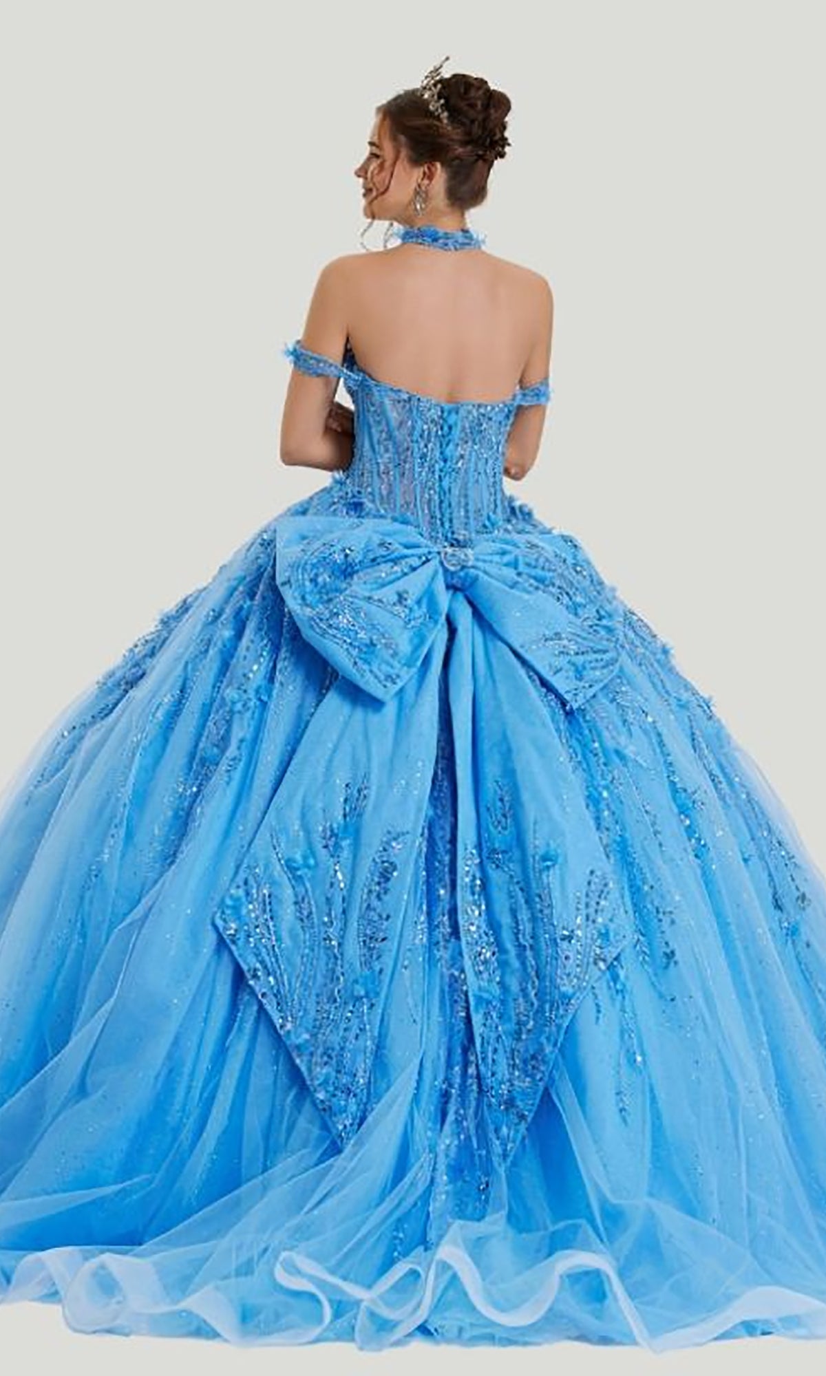 A woman stands with her back to the camera against a plain light background, wearing the Quinceanera Dress 26416 By Stella Couture—an ornate blue ball gown with a glitter tulle skirt, an embellished corset bodice, sparkling details, and a large bow.